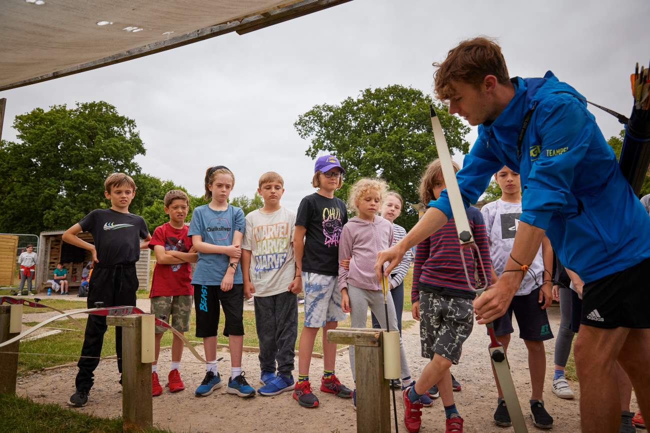 A coach demonstrating archery technique to a group of attentive children outdoors under a shelter, helping them discover the fundamentals of the sport.