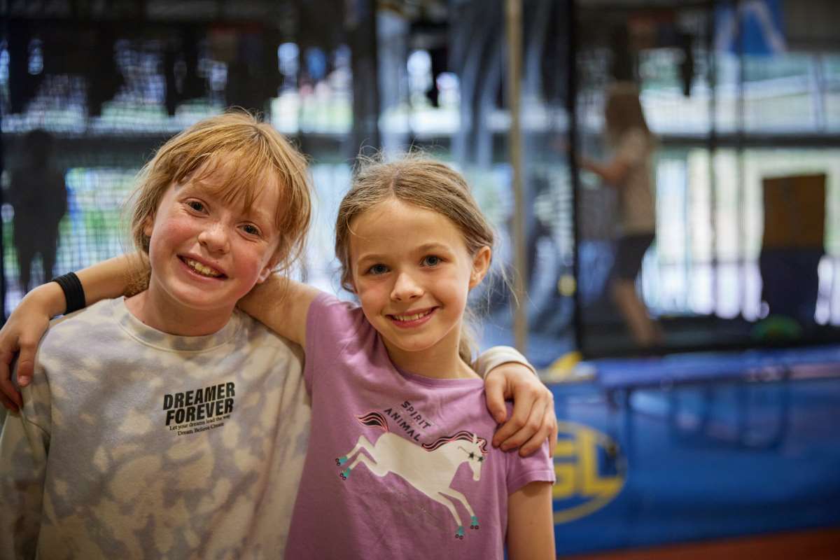 Two young girls smiling and embracing each other inside a gym, one wearing a "discover dreams" t-shirt and the other in a unicorn shirt.