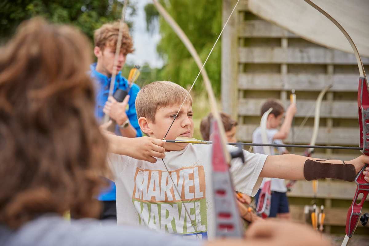 Young boy in a Marvel t-shirt concentrating while aiming with a bow and arrow at an archery range, surrounded by other participants, eager to discover his potential.