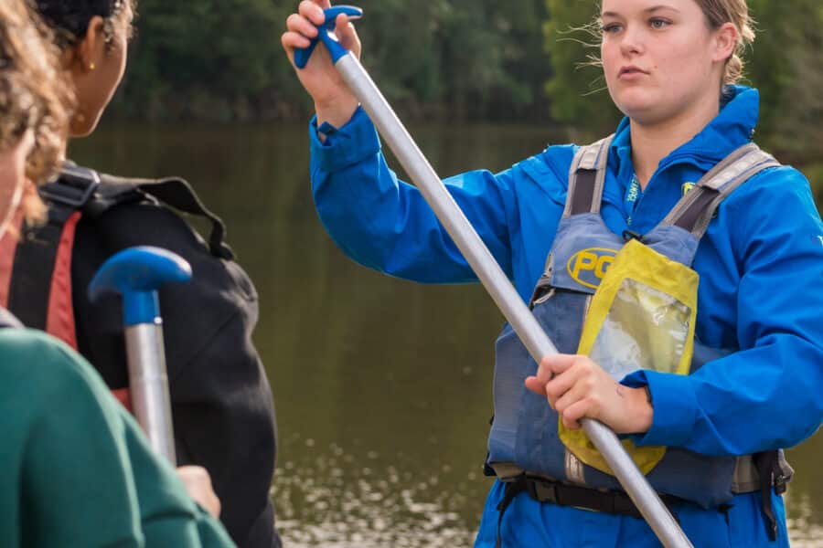 A woman in a blue jacket instructs two others on how to use a paddle on a riverbank.