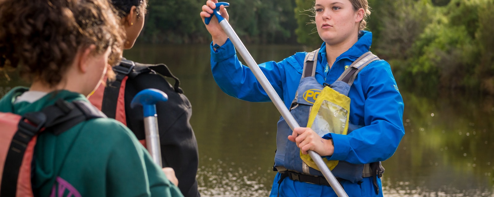 A woman in a blue jacket instructs two others on how to use a paddle on a riverbank.