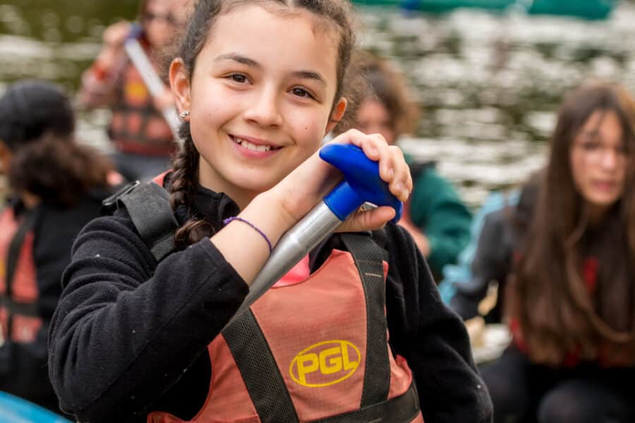 Young girl in a life vest smiling at the camera while holding an oar, with other children canoeing in the background.