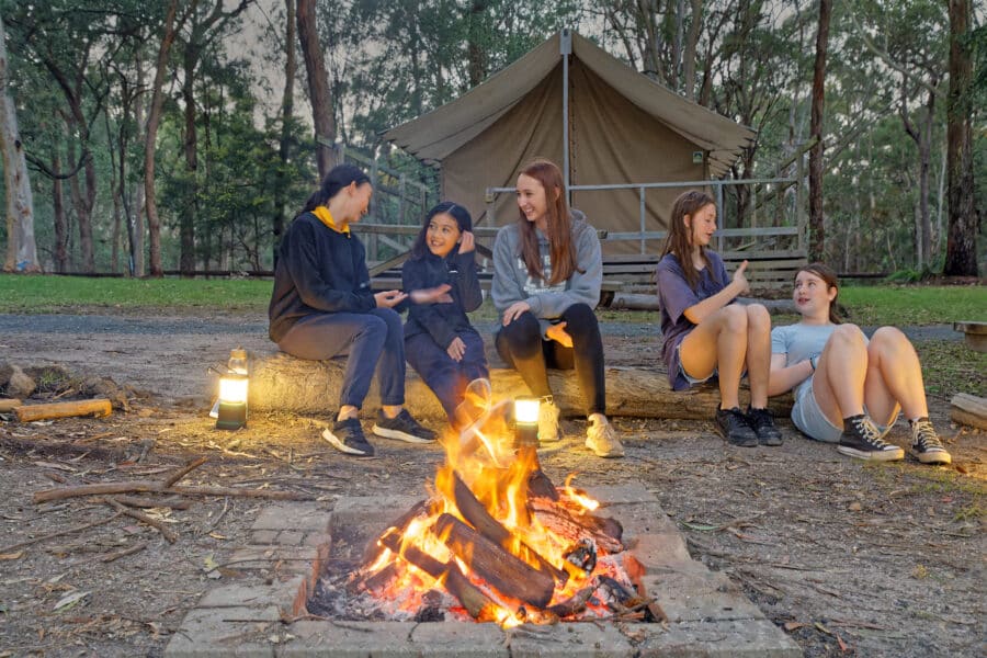 A group of five people sitting around a campfire in a forest setting near a tent, enjoying a conversation at dusk. two lanterns illuminate the area.