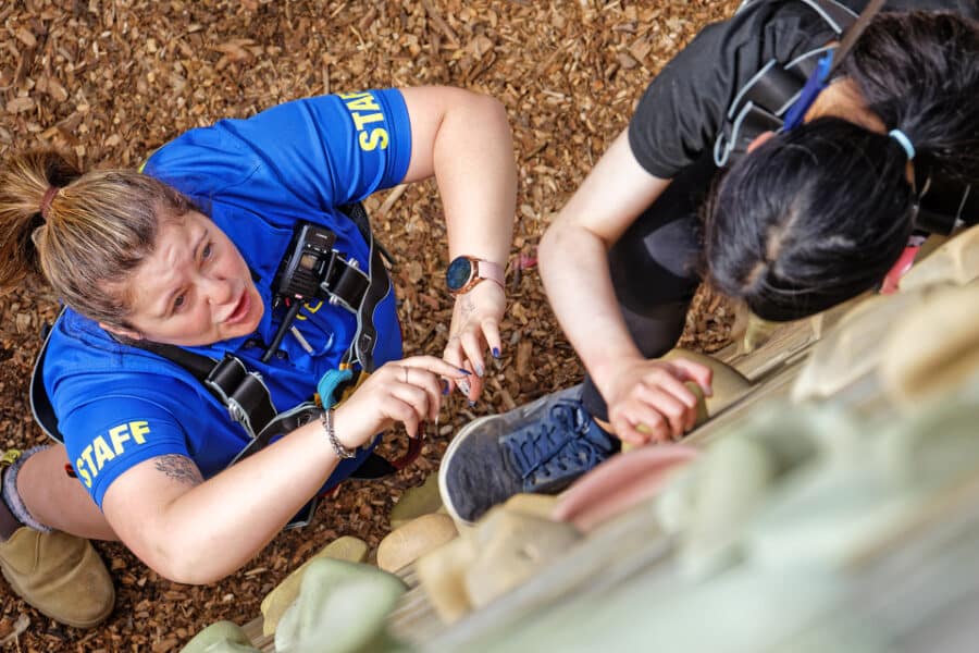 Staff member in a blue shirt assists a climber with a harness at an outdoor climbing wall, focusing intently on the climber's safety.