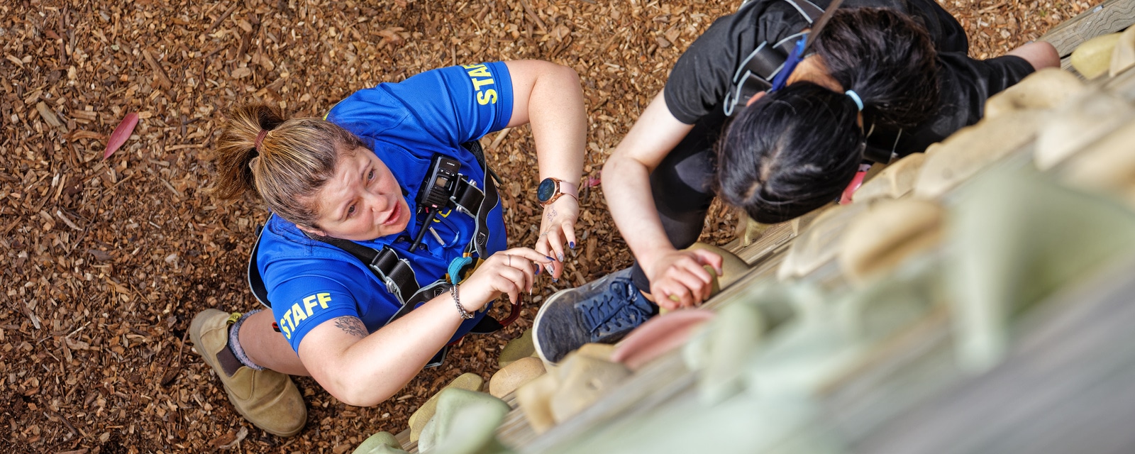 Staff member in a blue shirt assists a climber with a harness at an outdoor climbing wall, focusing intently on the climber's safety.