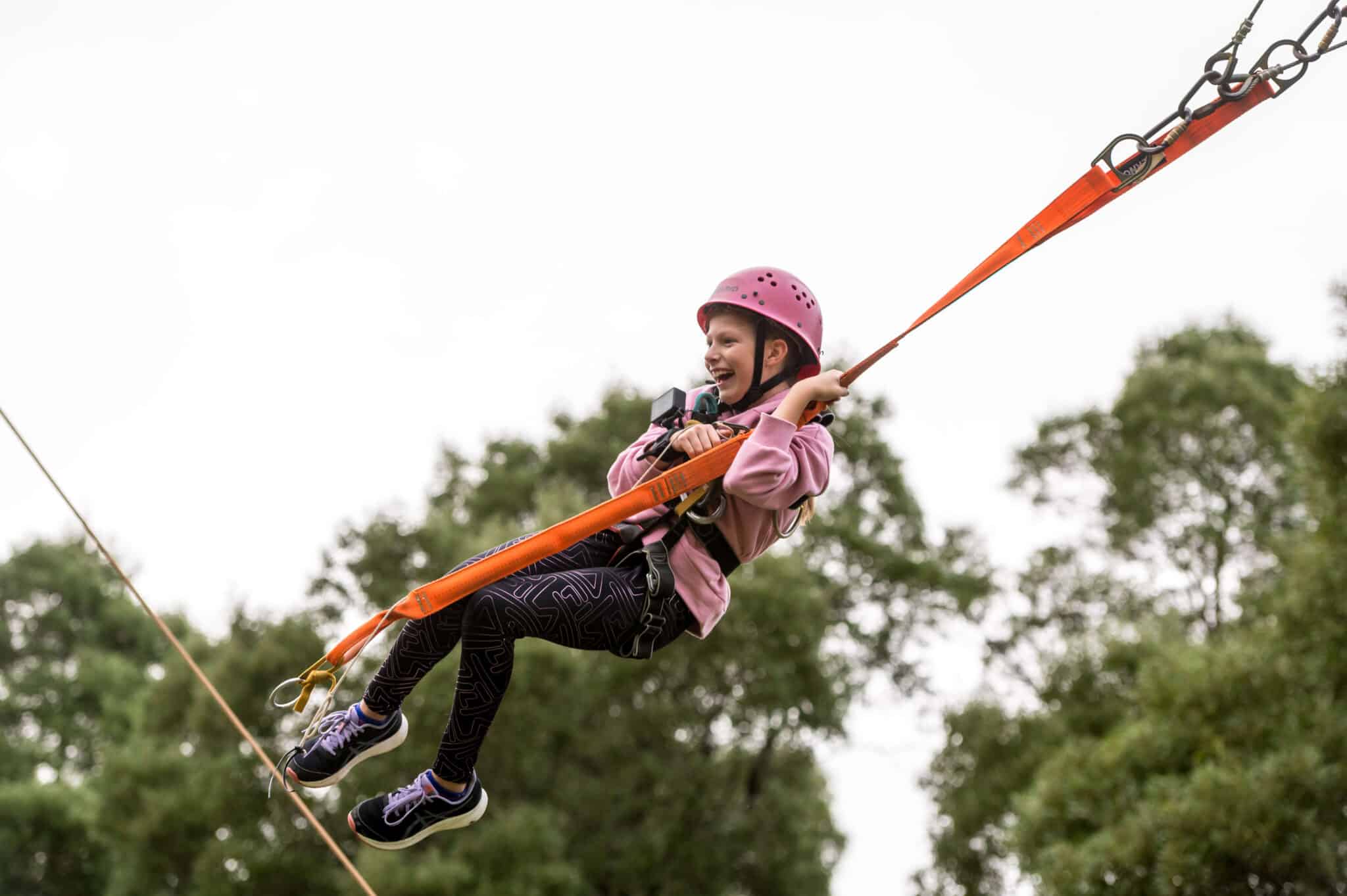 A young girl wearing a pink helmet and harness joyfully zip-lining against a backdrop of green trees.