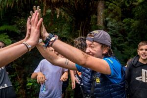 A woman in a blue shirt high-fiving someone at a group gathering in a lush forest setting, with smiling onlookers around, discovering the joy of communal activities.