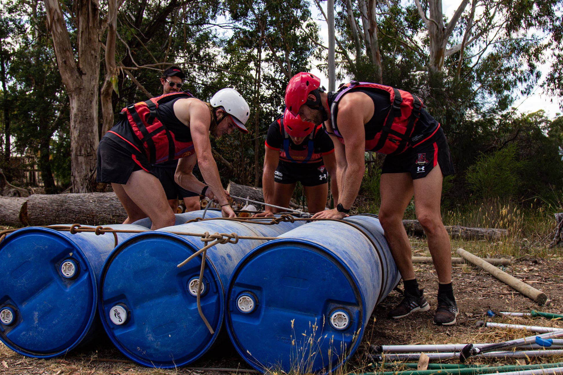 Three people in helmets and sports gear assembling a raft with blue barrels and wooden poles in a forested area.