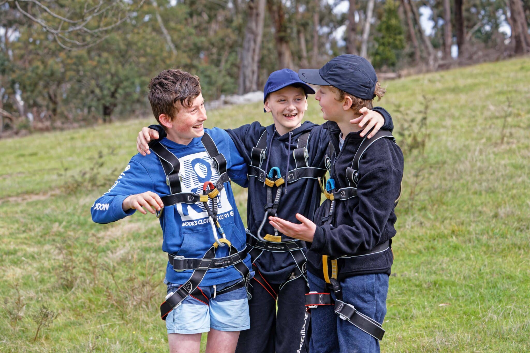 Three boys in outdoor clothing and harnesses laugh and interact joyfully in a grassy field.