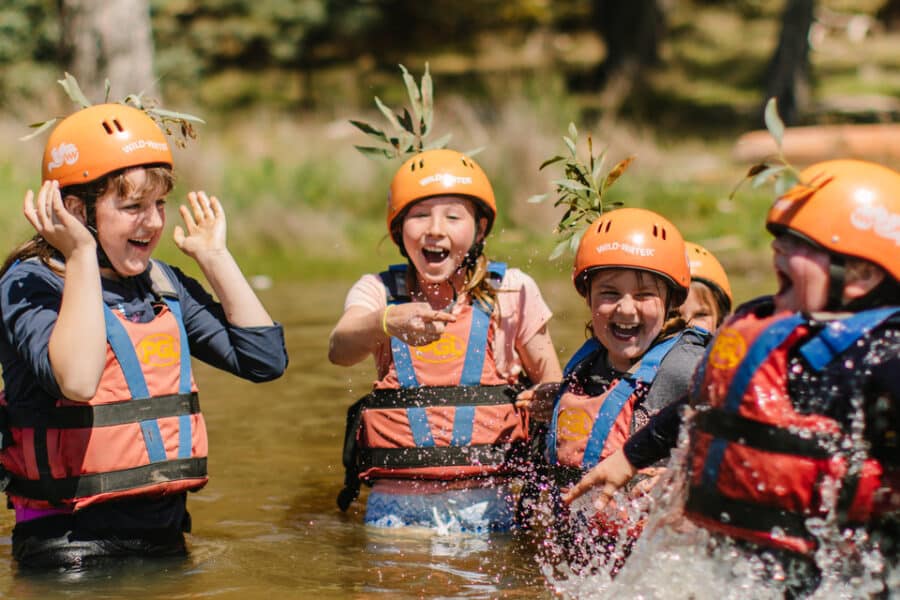 Group of children in safety helmets and life vests laughing and splashing in a river during an outdoor adventure activity.