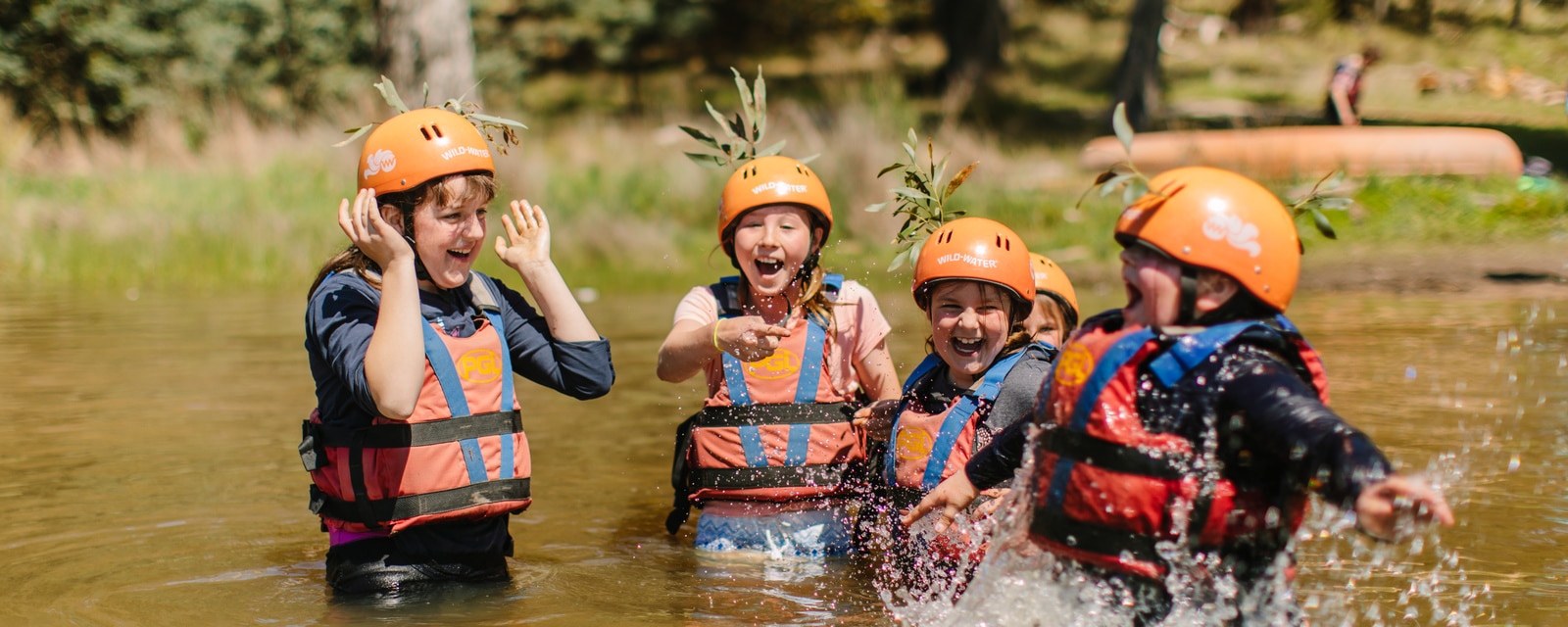 Group of children in safety helmets and life vests laughing and splashing in a river during an outdoor adventure activity.