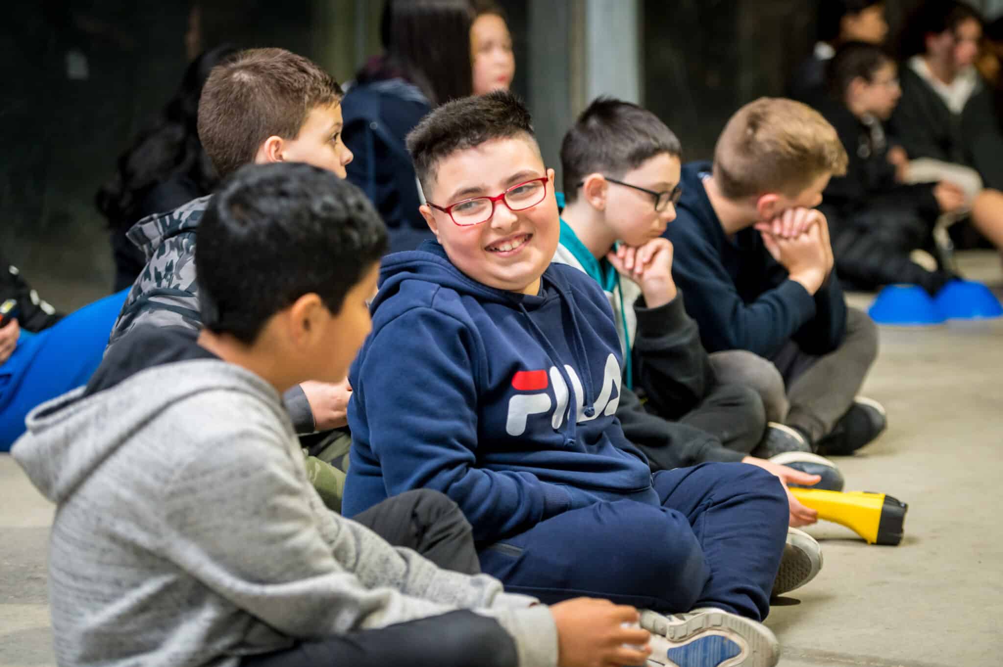 Group of children sitting on the floor in a semi-circle, smiling and listening to a speaker at a school event.