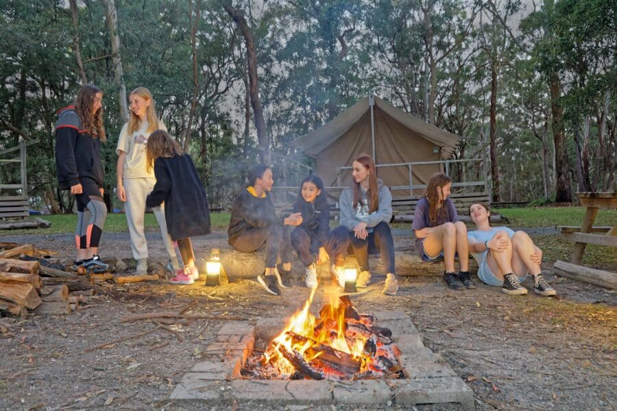 Group of teens enjoying a conversation around a campfire with a tent in the background at dusk.