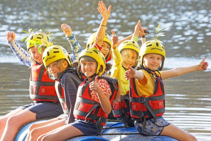 A group of six children wearing yellow helmets and red life jackets joyfully pose on a raft, raising their hands and smiling, water in the background.