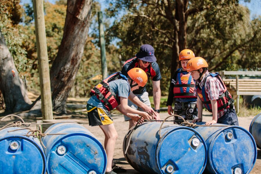 Three people in helmets and life vests assemble a raft using blue barrels in a sunny outdoor setting.