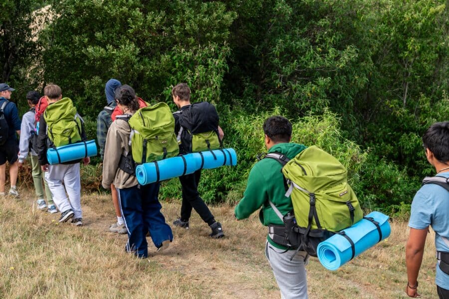 A group of hikers with green backpacks and blue rolled mats walk in a line along a grassy trail beside dense green trees.