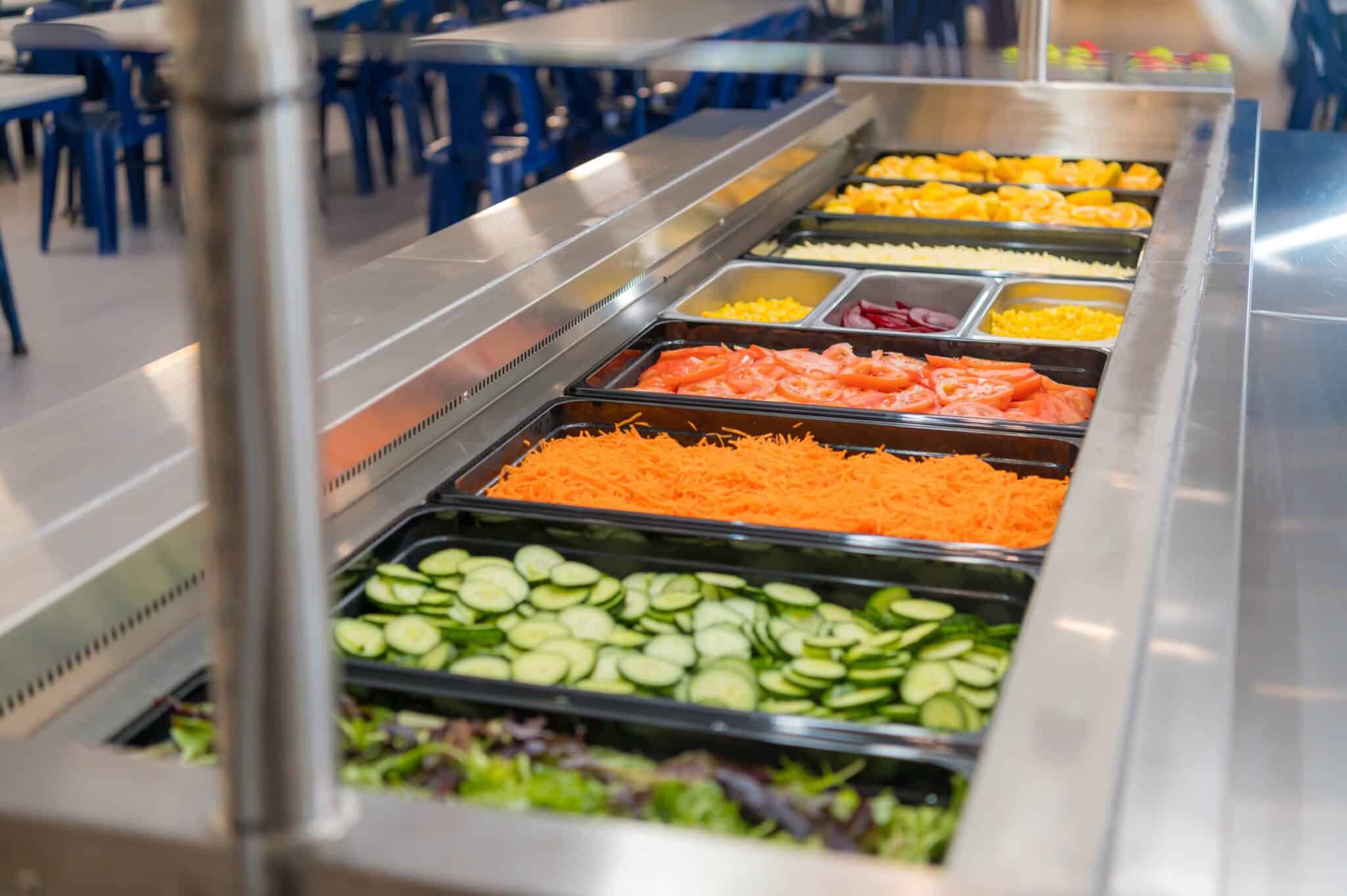 A salad bar uniting trays of fresh cucumbers, carrots, tomatoes, peppers, and other vegetables in a cafeteria.