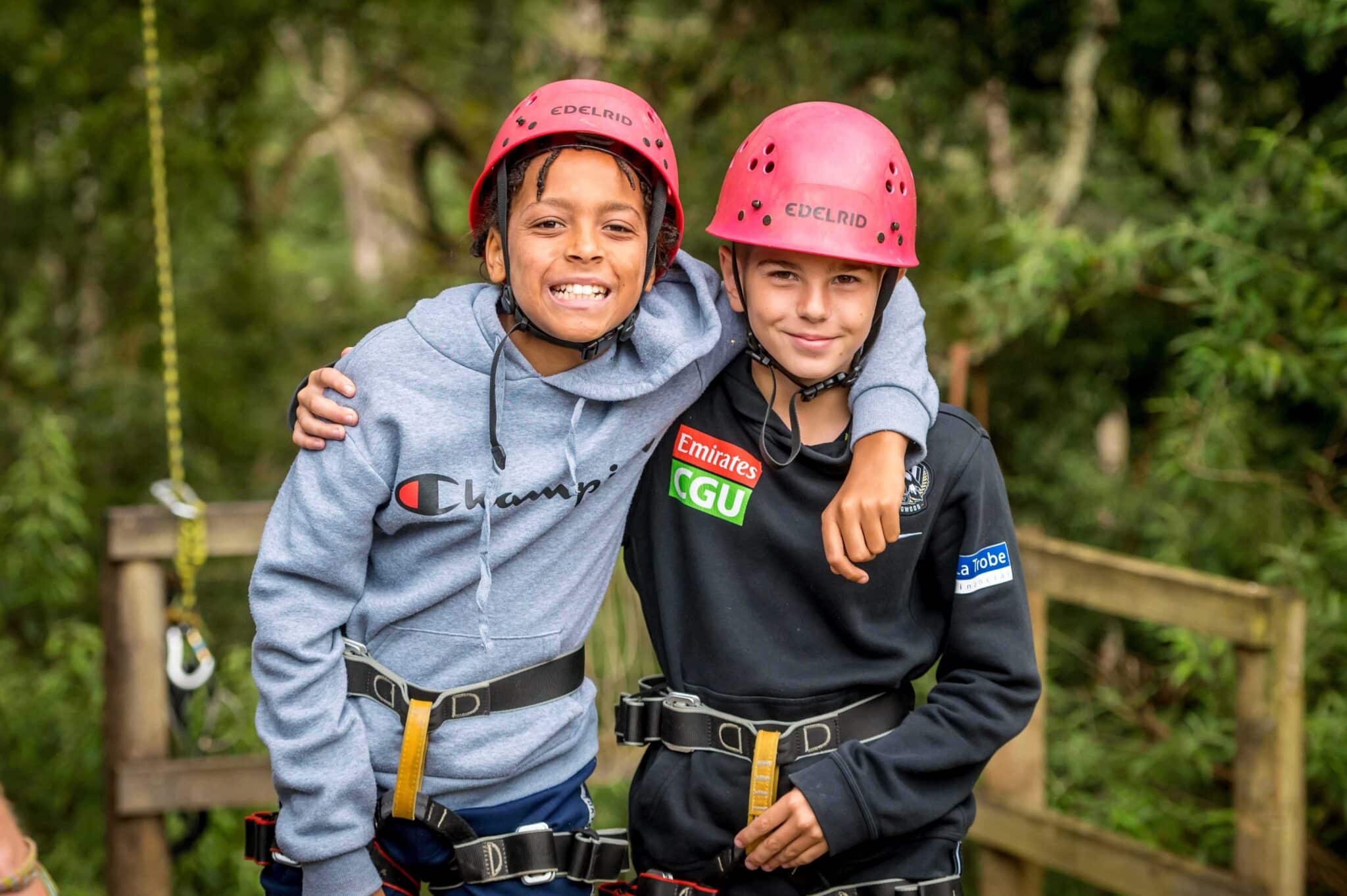 Two children wearing helmets and harnesses unite in an embrace, smiling as they stand outdoors in a wooded area.