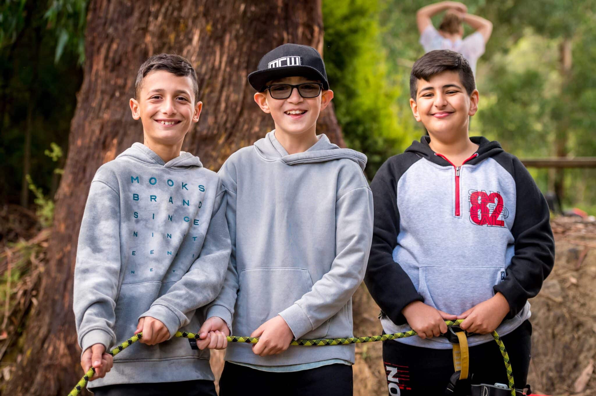 Three boys unite smiling outdoors, two gripping a rope; one wearing glasses and a cap, and a blurred person visible in the background.