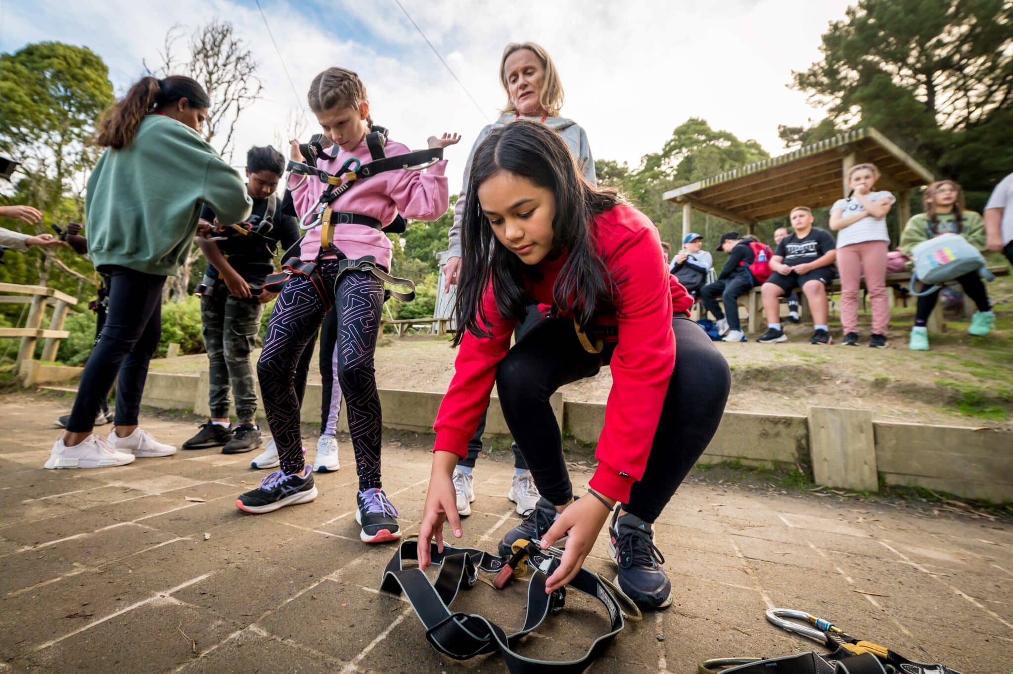 A girl secures her shoes while a group of young people unite to prepare for an outdoor activity, with an instructor assisting in the background.