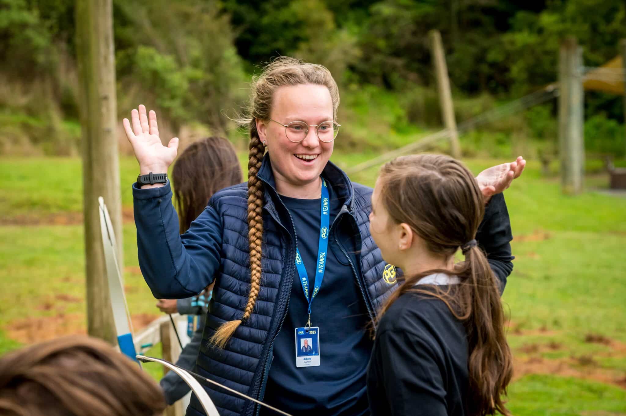 A smiling woman with glasses wearing a lanyard talks animatedly with a young girl in a park, gesturing with her hands to unite their ideas.