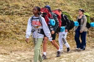 A group of teenagers hiking on a grassy trail, discovering nature as they carry backpacks and rolled-up sleeping mats.