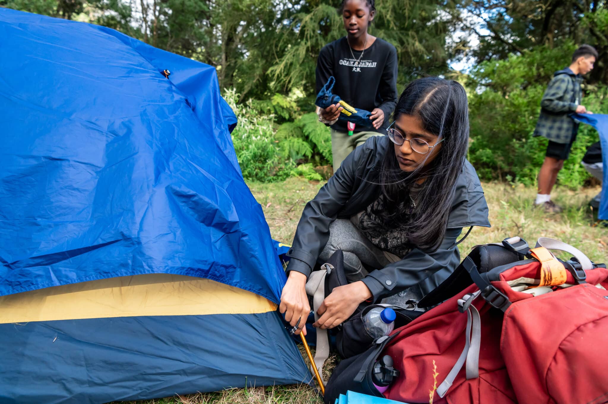 A woman in glasses pitches a blue tent in a forested area, with another person standing nearby and gear scattered around, ready to discover the wilderness.