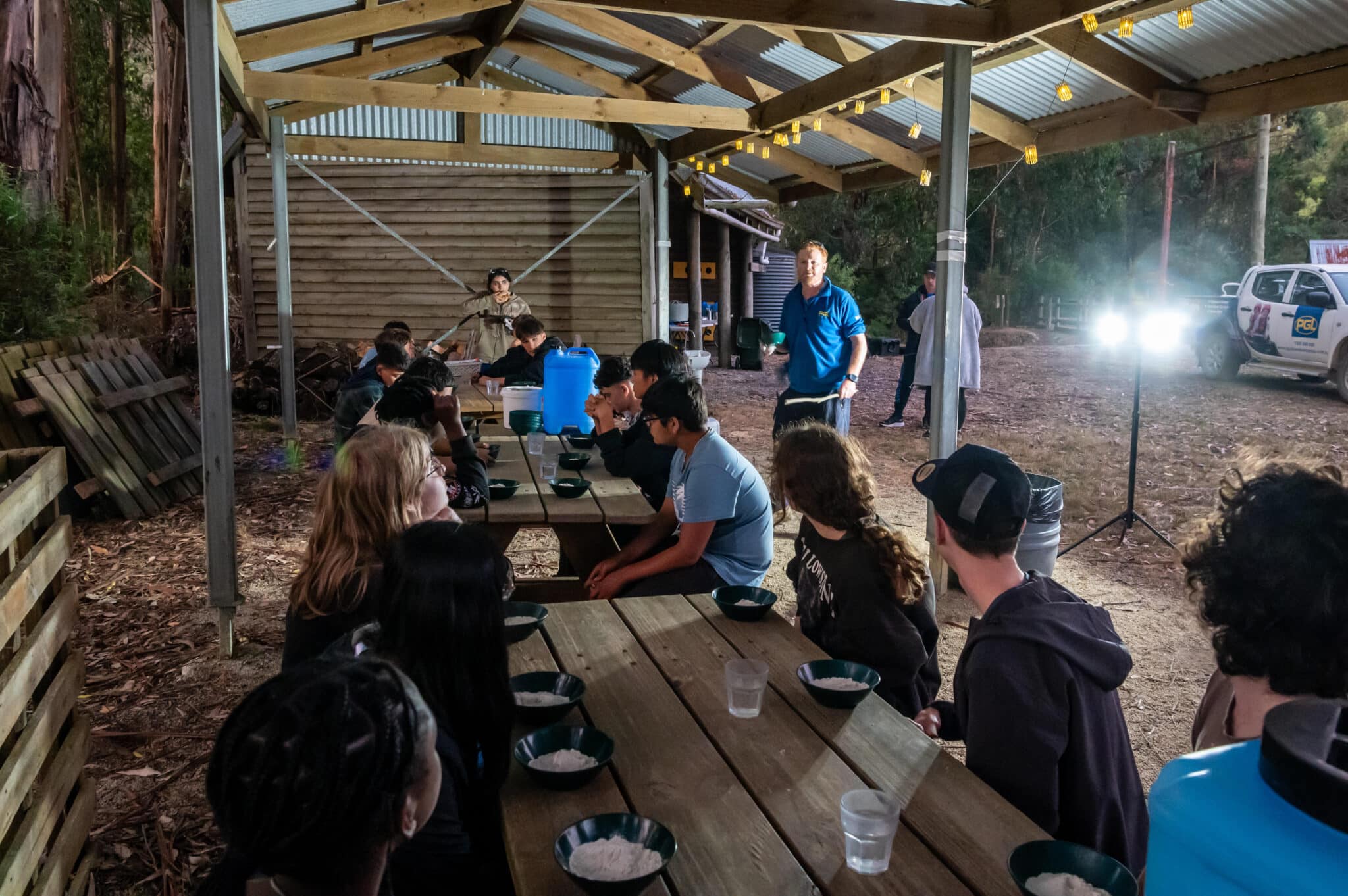People gathered at an outdoor covered seating area with wooden tables, engaging in a meeting during evening hours, illuminated by artificial lights to discover new ideas.