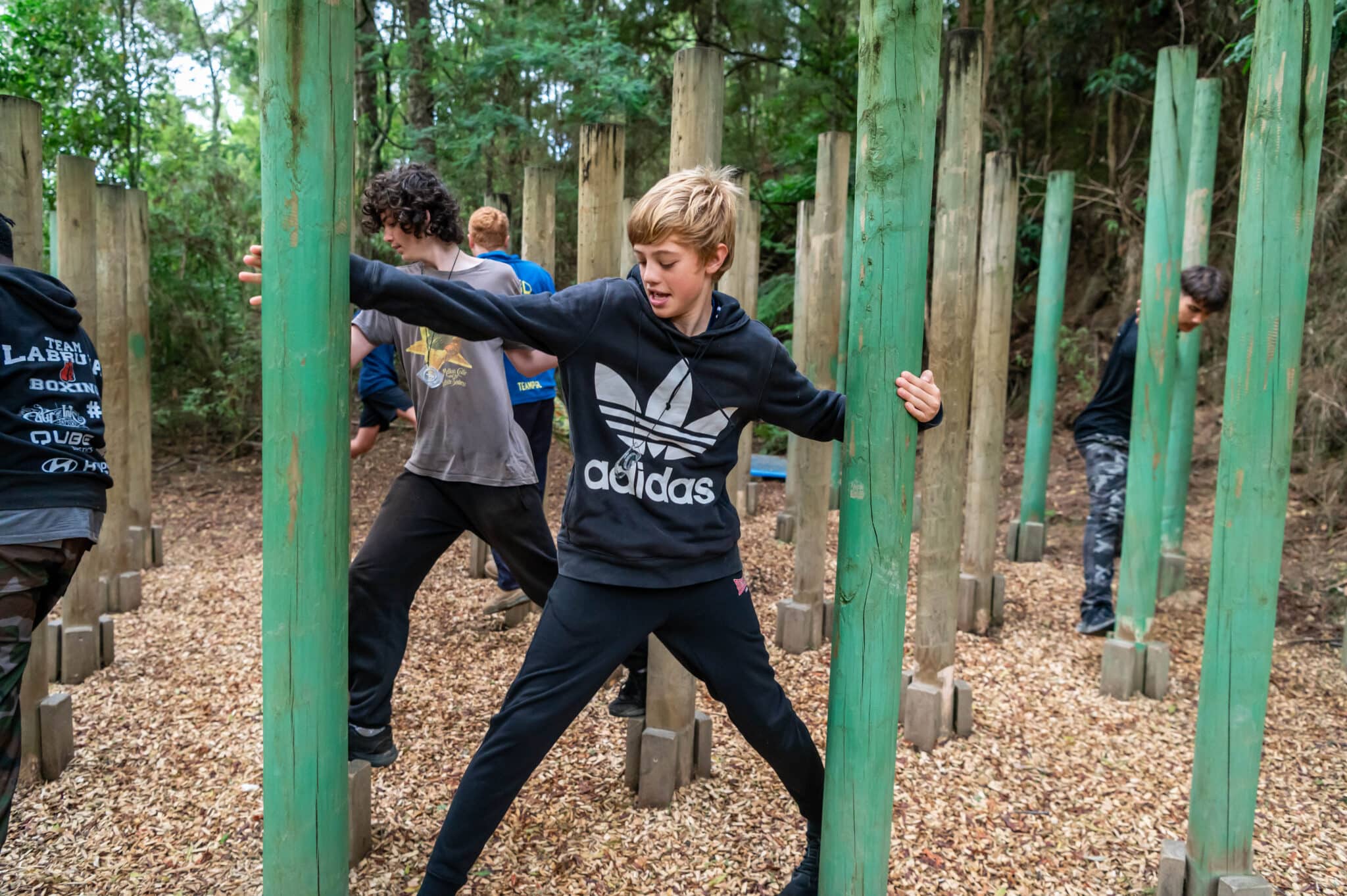 A boy in a black adidas sweatshirt discovers his way through a wooden pole obstacle course in a forest setting, showing concentration and movement.