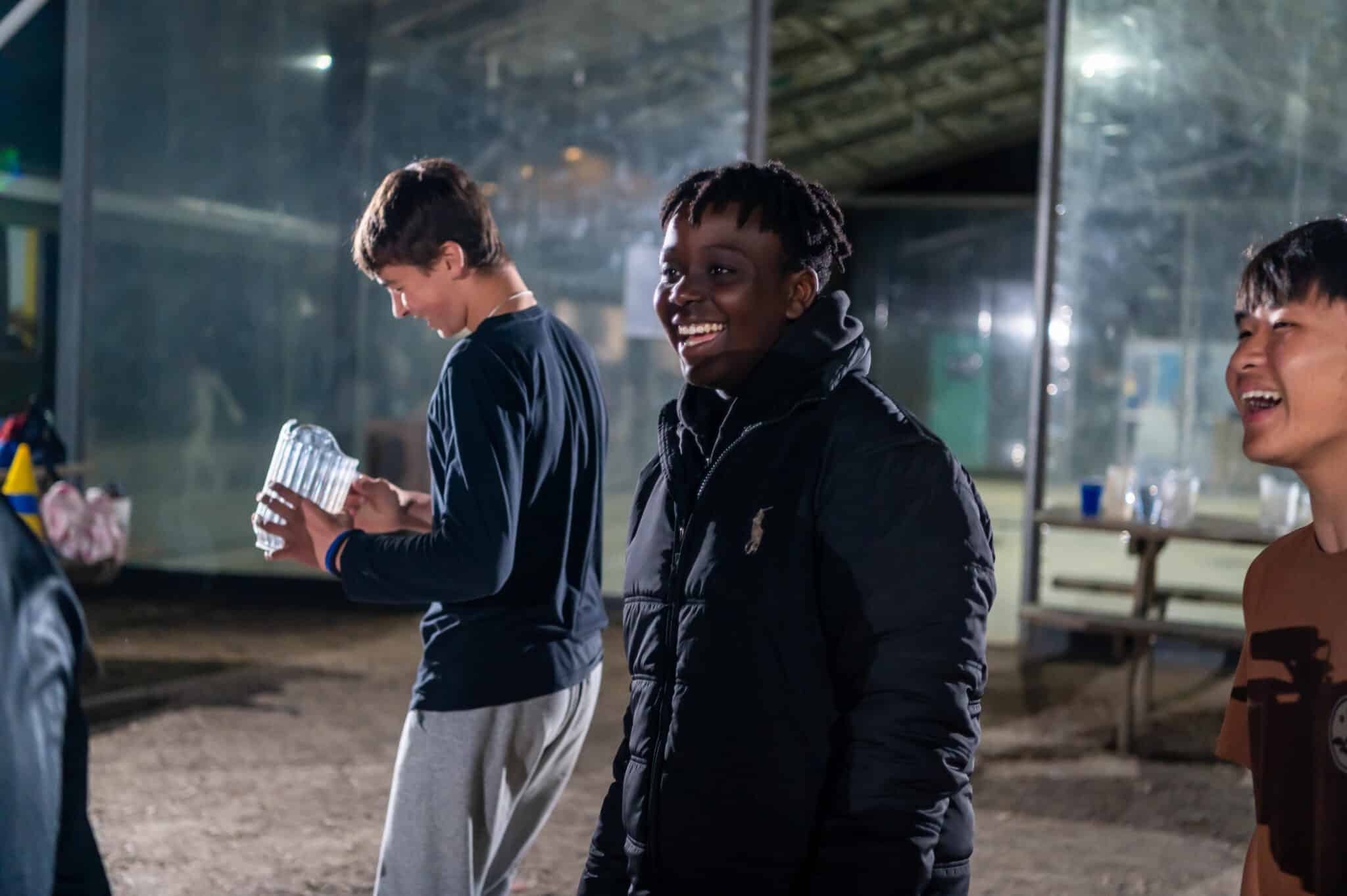 Three young men smiling and laughing at night, one holding a water bottle, with a blurred background suggesting they are discovering the vibes of a busy outdoor setting.