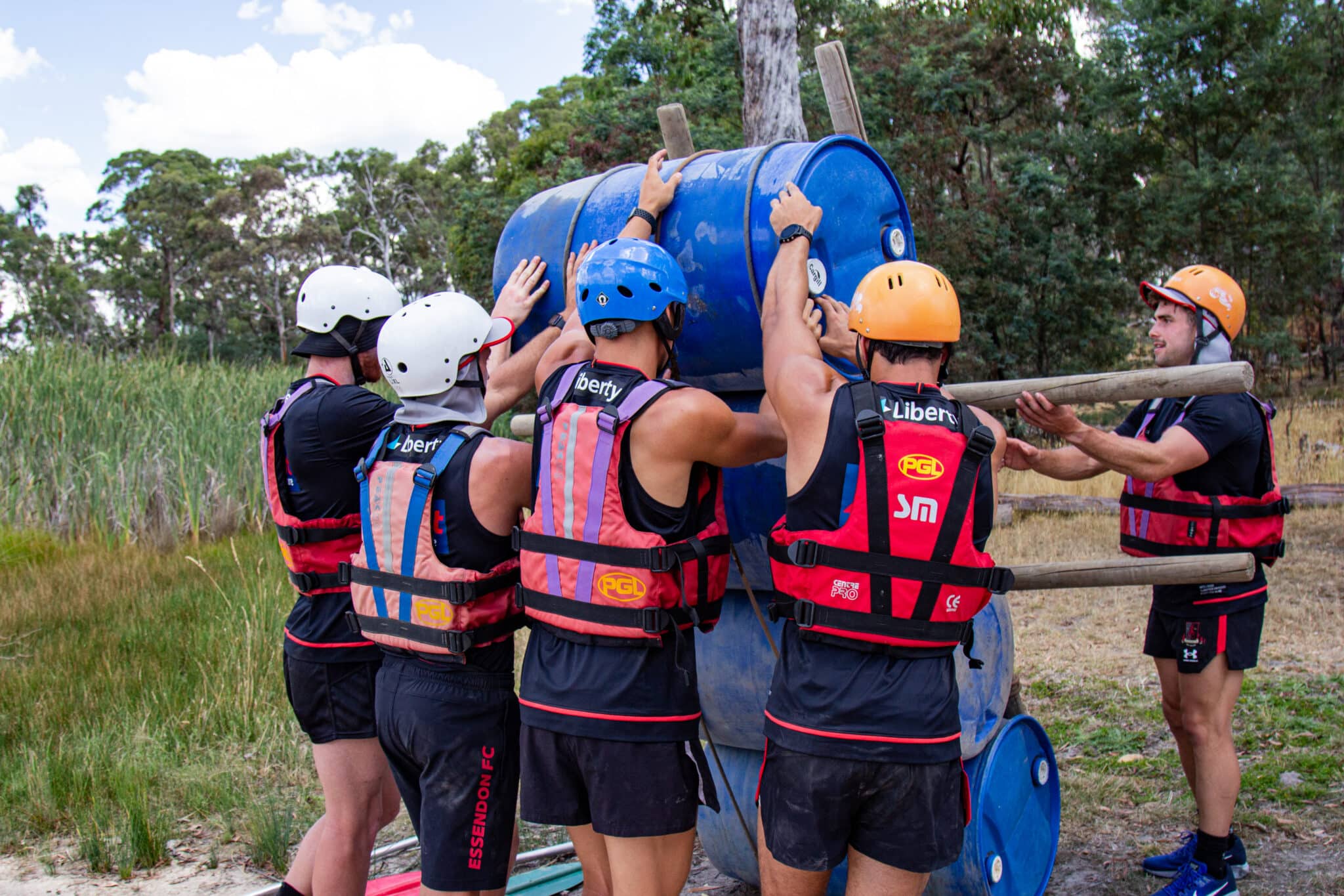 A group of people wearing life vests and helmets work together to build a structure using blue barrels and wooden poles outdoors near a forested area, showcasing the spirit of teamwork common in group camps.