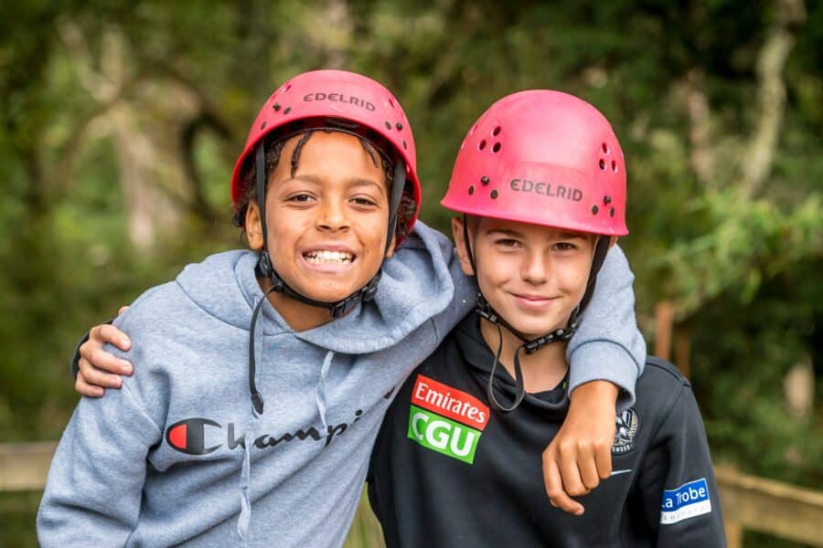 Two children wearing safety helmets and sportswear smile with their arms around each other, standing outdoors with a green, wooded background, as if enjoying the camaraderie of group camps.