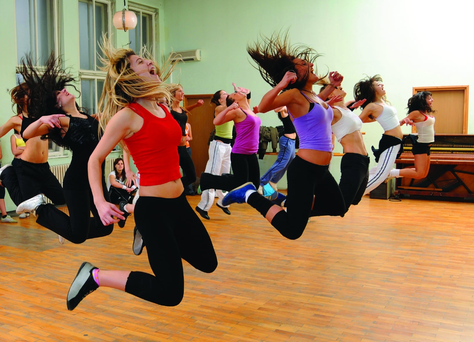 A group of people in athletic clothing are mid-air, performing a jump during a dance class in a studio with wooden flooring, reminiscent of the spirited energy found at group camps.
