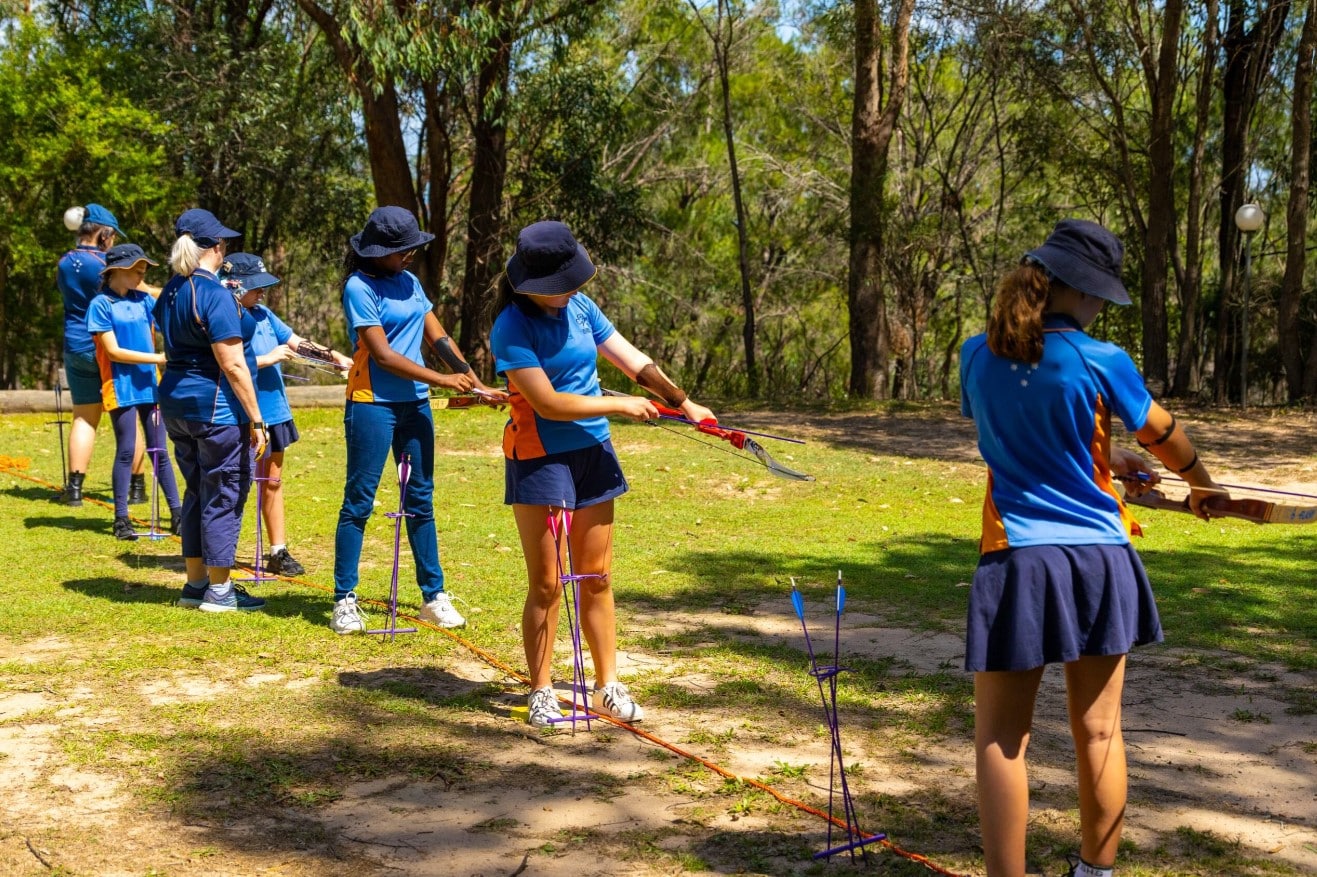 A group of people in blue uniforms practice archery outdoors in a wooded area, with targets placed in front of them as part of their faith camp activities.