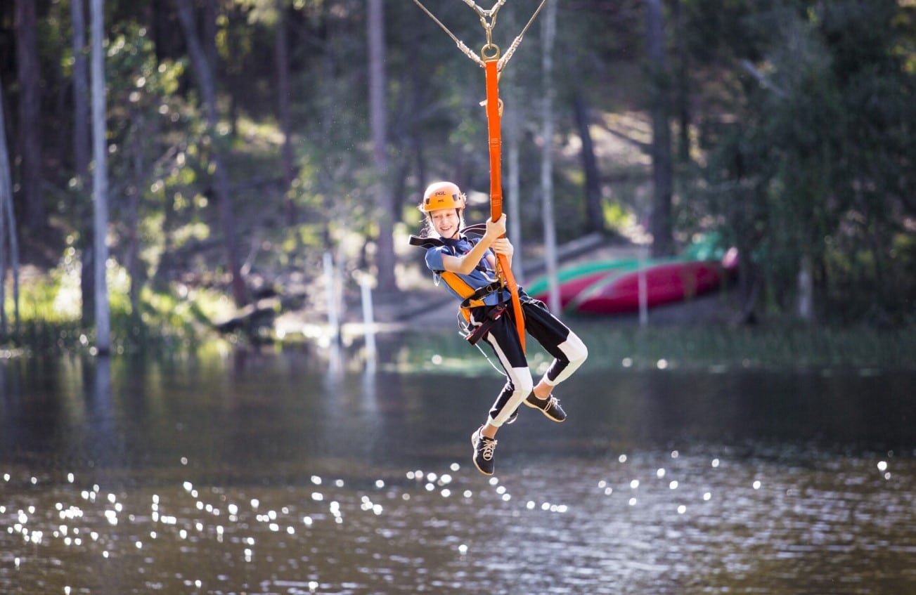 A person wearing a helmet and harness rides a zipline over a body of water, with trees and a canoe visible in the background, evoking the adventurous spirit often found at church camps.