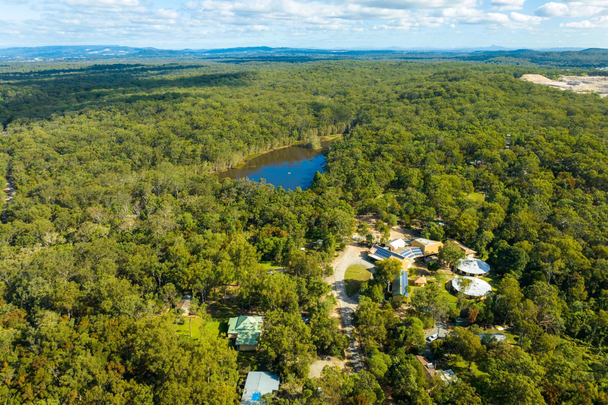 Aerial view of a lush, expansive forest with a small lake and scattered buildings in a clearing under a partly cloudy sky.