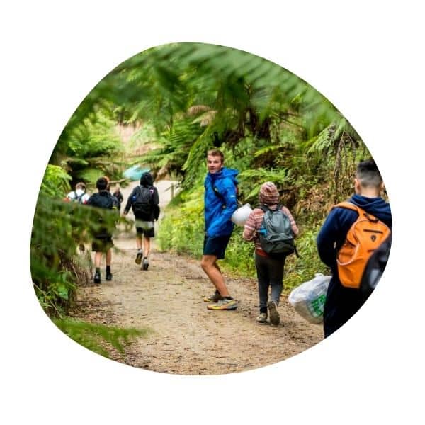 A group of people wearing backpacks walk along a forest trail. One person looks back toward the camera.
