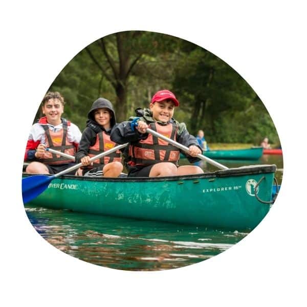 Three kids in life jackets paddle a green canoe on calm water with trees in the background.
