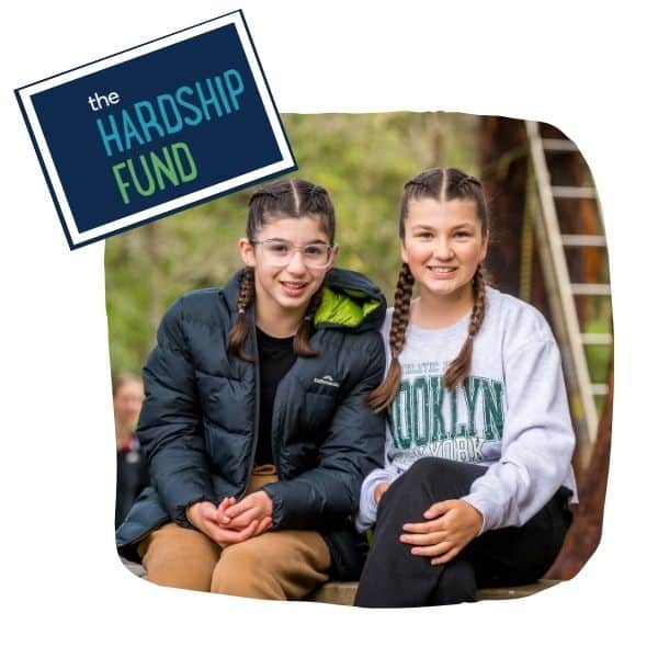 Two smiling girls with braided hair sit outdoors. One wears glasses and a black jacket, the other a gray Brooklyn sweatshirt. A "The Hardship Fund" sign is in the corner.