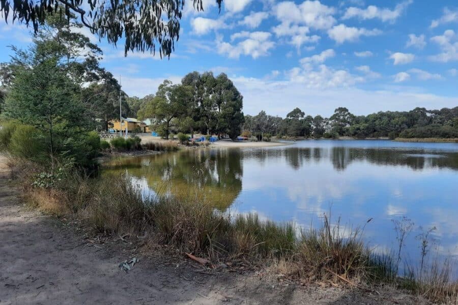 A serene lake surrounded by trees with a clear sky and scattered clouds. A few buildings and trees are reflected on the water's surface.