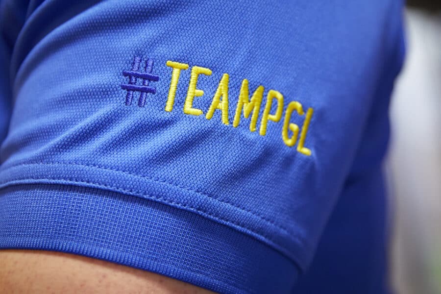 Close-up of a blue shirt sleeve with the yellow embroidered text "#TEAMPGL" and part of a PGL logo visible in the background.