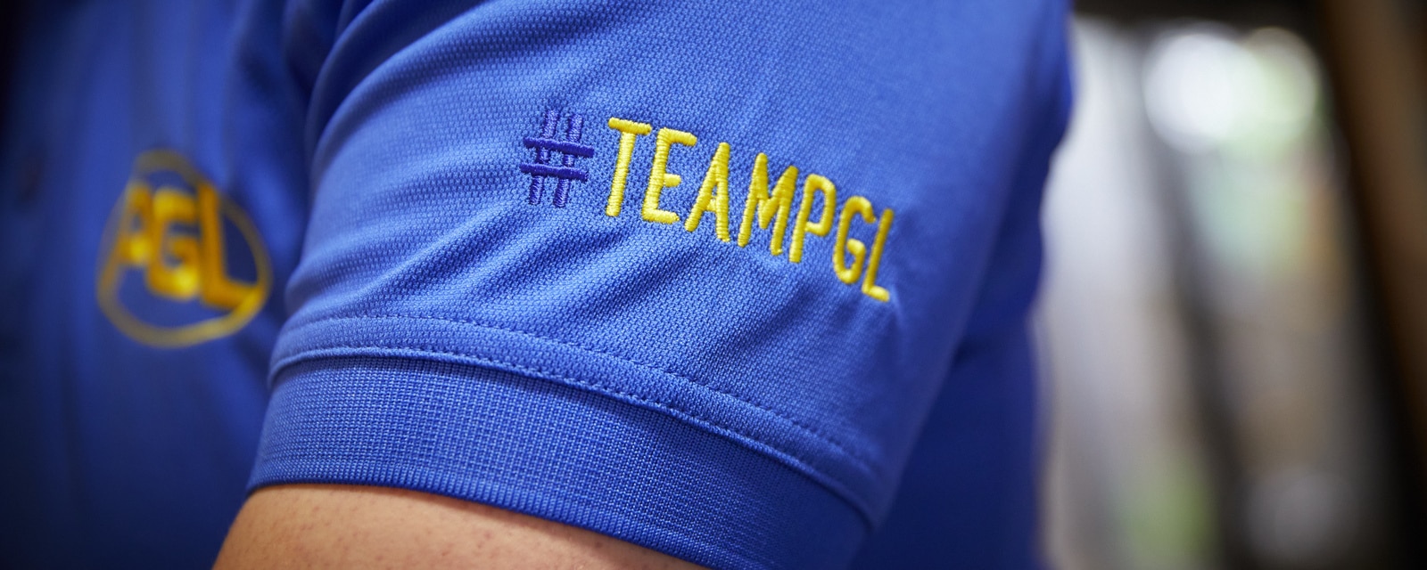 Close-up of a blue shirt sleeve with the yellow embroidered text "#TEAMPGL" and part of a PGL logo visible in the background.