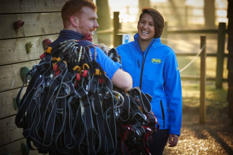 Two people in blue jackets are standing outdoors; one is carrying several climbing harnesses, and the other is smiling. They are near a wooden climbing wall and a wooden fence.