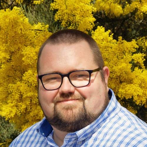 A man with glasses and a beard stands in front of bright yellow flowering bushes, wearing a blue and white checked shirt.
