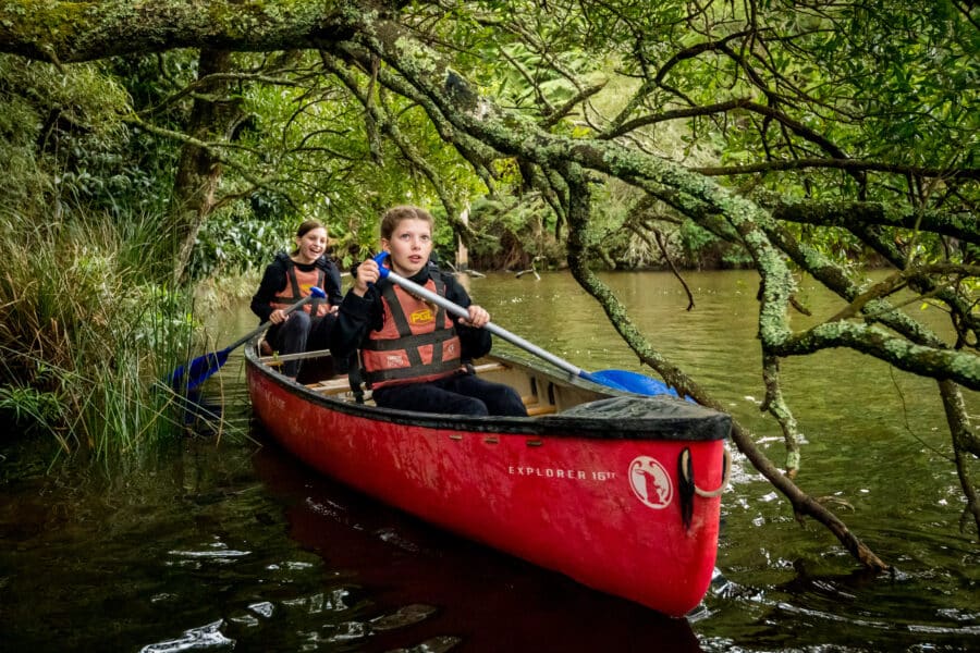 Two people wearing life jackets paddle a red canoe under low-hanging tree branches on a calm, narrow river surrounded by dense greenery.
