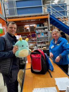 Two people stand at a table in a warehouse; one holds a green plush toy, while the other sorts items near a red and blue backpack. Shelves with boxes and supplies are in the background.