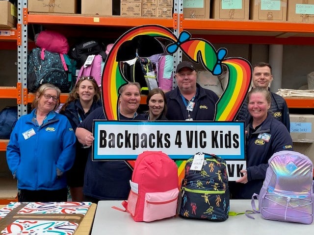 Seven people stand in a warehouse, holding a "Backpacks 4 VIC Kids" sign, with colorful backpacks displayed on a table in front of them. Shelves of bags are seen in the background.